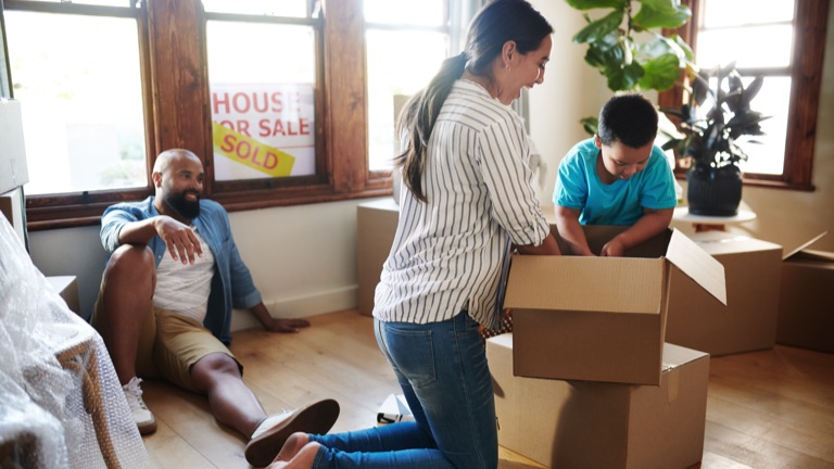 A family packing moving boxes in a living room, a sold sign hanging in the window behind them