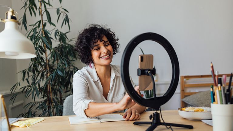 Woman sitting at an office desk with recording a video on her cell phone with a ring light.