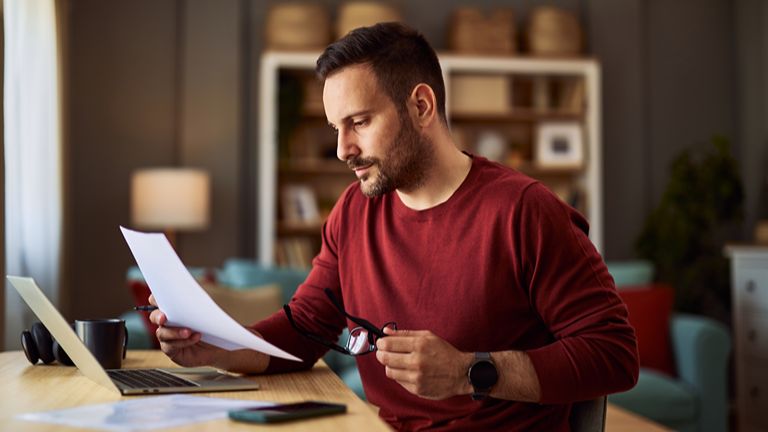 A busy young adult male freelancer review a piece of paper while working on a project on a laptop
